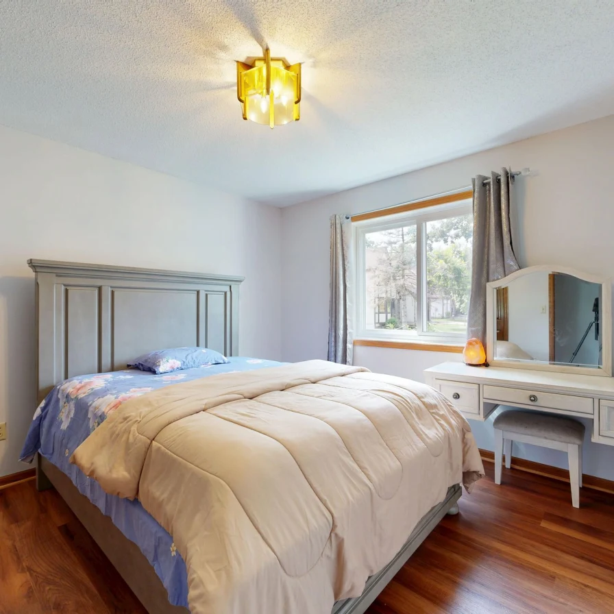 Dormitorio con una cama de edredón beige, una ventana con árboles y un tocador con espejo