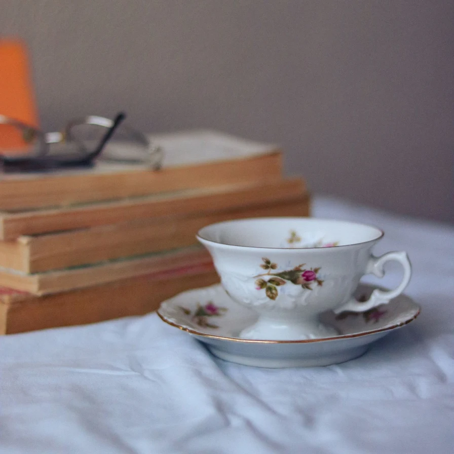 Taza de porcelana floreada sobre una cama, con libros y unas gafas desenfocados detrás