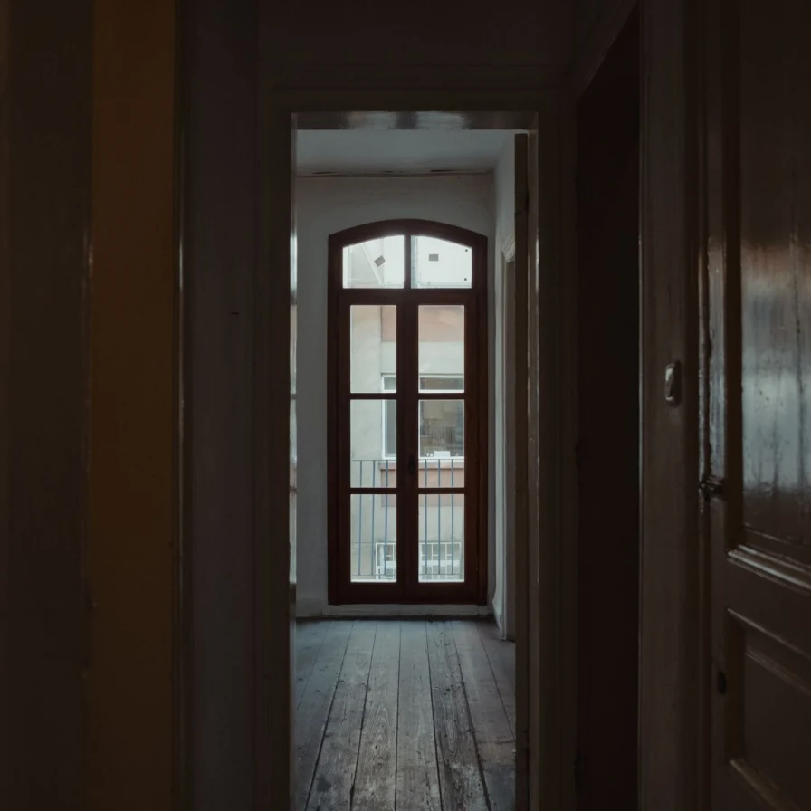 Pasillo de una casa antigua con puertas de madera y suelo de tablones hacia una puerta acristalada que da a un balcón