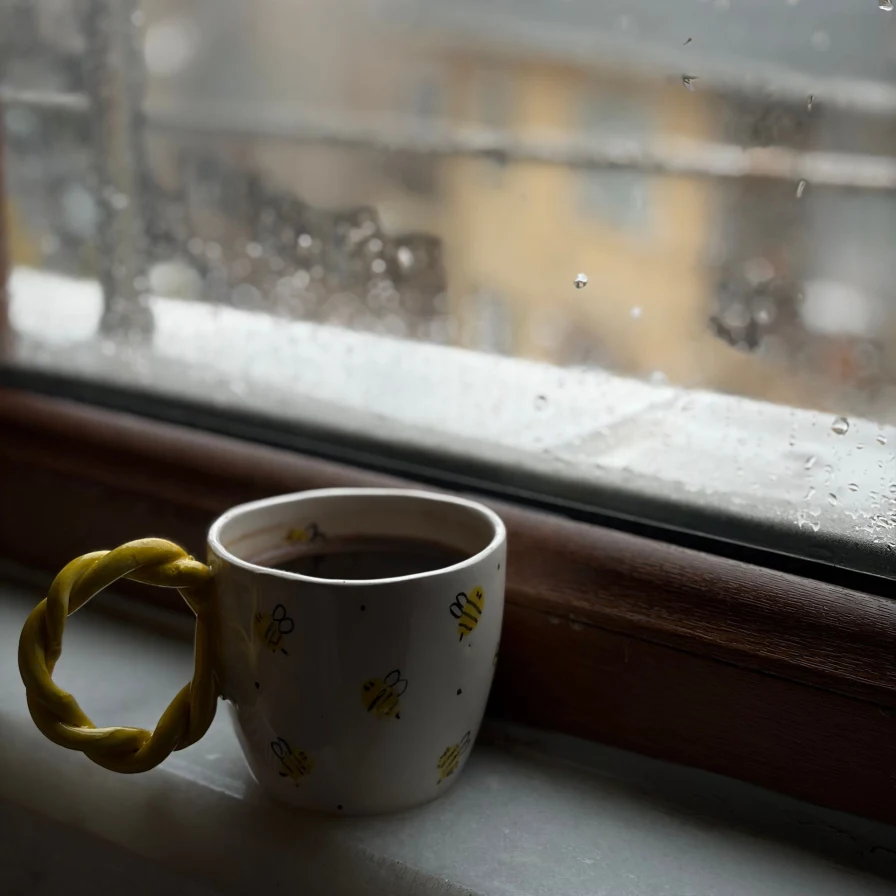 Taza de cerámica con dibujos de abejas sobre el alféizar de una ventana con gotas de lluvia
