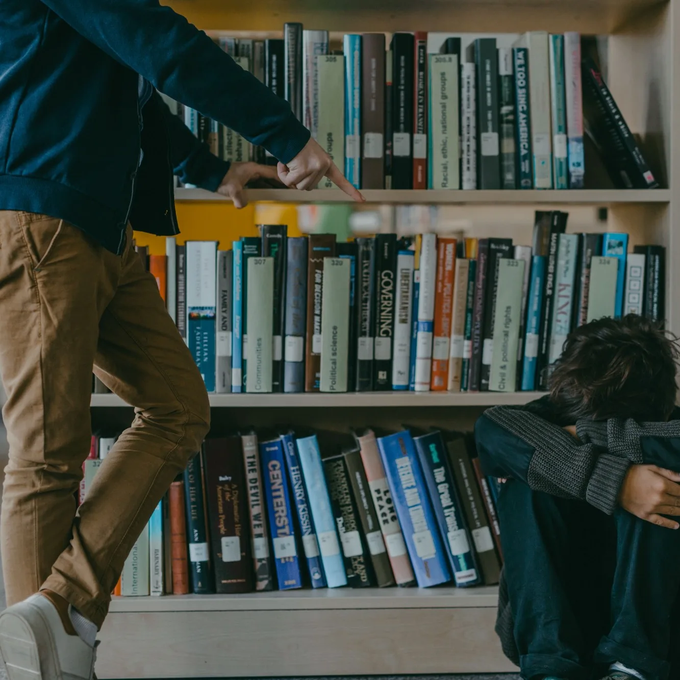 Fotografía: un niño encogido en el suelo de una biblioteca mientras otra persona le señala con el dedo